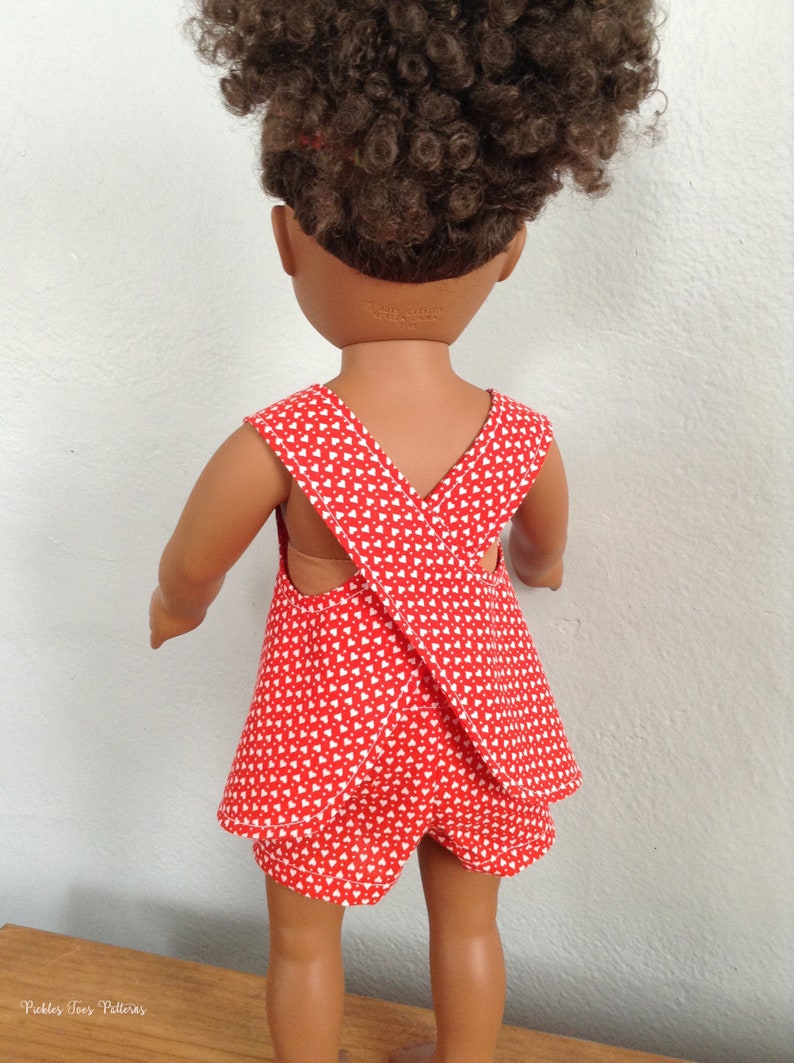 Puede incluir: Un vestido de mu&ntilde;eca rojo y blanco con una correa cruzada en la espalda. El vestido tiene un patr&oacute;n de corazones blancos sobre un fondo rojo. El vestido lo lleva una mu&ntilde;eca con el pelo rizado marr&oacute;n.