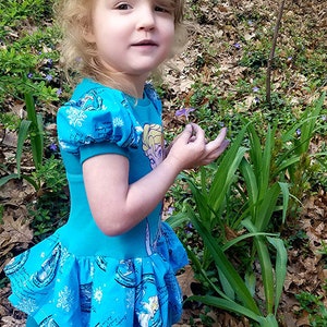 May include: A young girl wearing a blue dress with a Frozen print. The dress has a ruffled bottom and puffed sleeves. The girl is holding a purple flower in her hand. The dress is made by Pickles Toes Patterns.