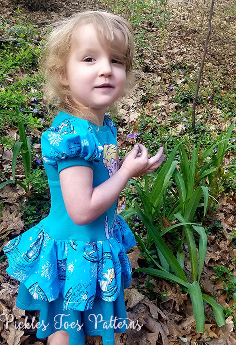 May include: A young girl wearing a blue dress with a Frozen print. The dress has a ruffled bottom and puffed sleeves. The girl is holding a purple flower in her hand. The dress is made by Pickles Toes Patterns.