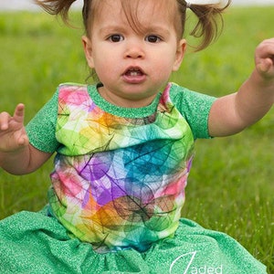 May include: A young child wearing a green and multi-colored leaf print shirt with a green ruffled skirt. The child has their hair pulled back in pigtails.