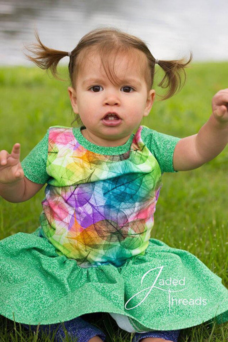 May include: A young child wearing a green and multi-colored leaf print shirt with a green ruffled skirt. The child has their hair pulled back in pigtails.