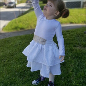 May include: A young girl dressed as a character from Star Wars, wearing a white dress with a silver belt and holding a blue lightsaber. The girl is standing on a grassy lawn.