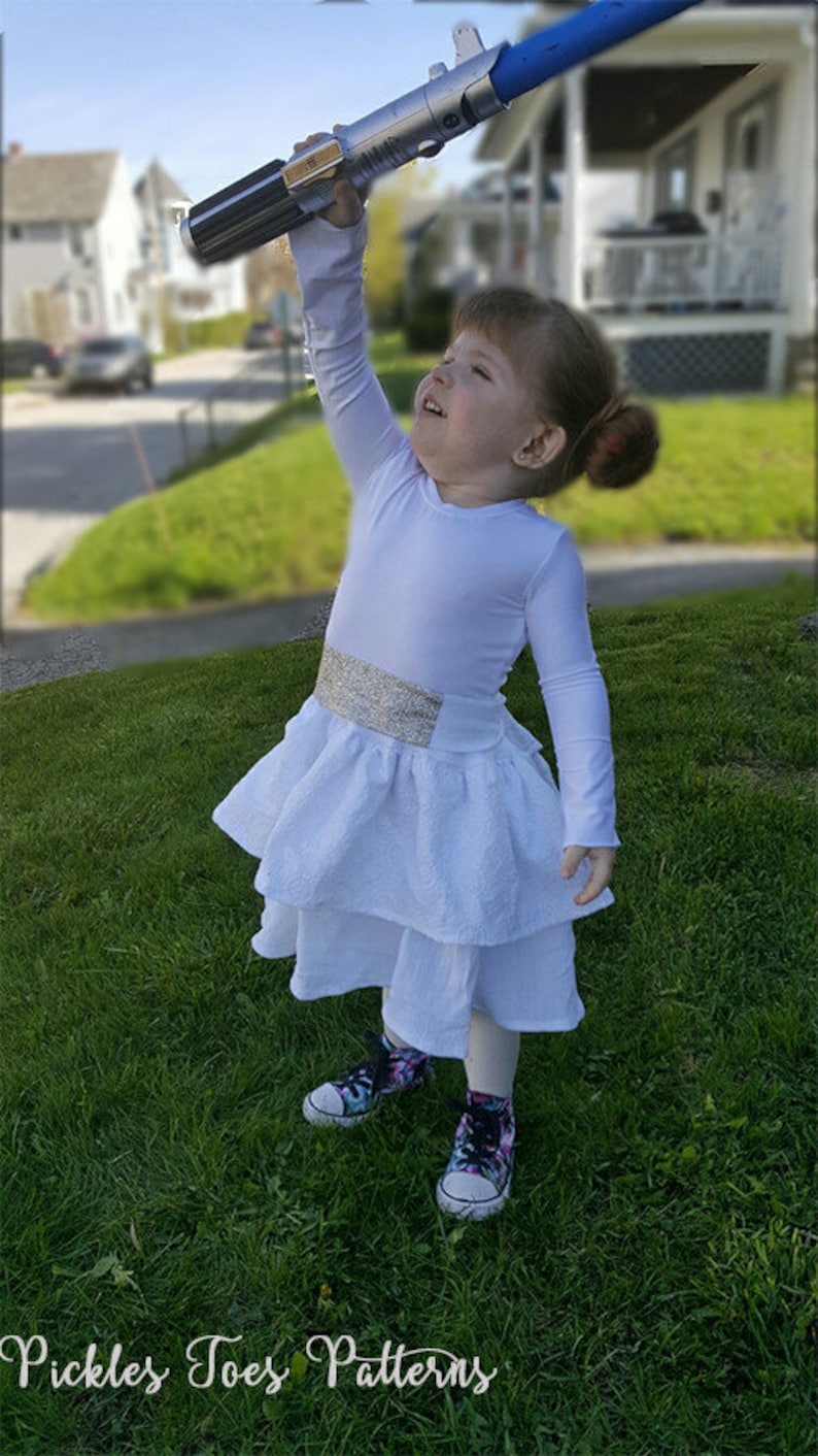 May include: A young girl dressed as a character from Star Wars, wearing a white dress with a silver belt and holding a blue lightsaber. The girl is standing on a grassy lawn.