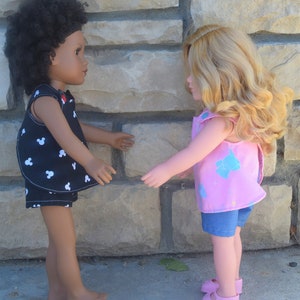 Puede incluir: Dos mu&ntilde;ecas con atuendos de verano se miran de frente. La mu&ntilde;eca de la izquierda lleva un vestido de estampado de Mickey Mouse en blanco y negro y pantalones cortos de mezclilla. La mu&ntilde;eca de la derecha lleva un vestido rosa sin mangas con estampado azul y blanco y pantalones cortos de mezclilla. Ambas mu&ntilde;ecas llevan zapatos rosas.