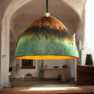 May include: A pendant light fixture with a dome-shaped shade. The shade transitions from orange to black to green, with a gold-colored interior. The light hangs from a black cord with a brass-colored fitting. The fixture is suspended in a room with arched ceilings.