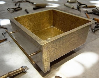 May include: A square, hammered brass kitchen sink with a single, horizontal bar handle. The sink is sitting on a concrete surface.