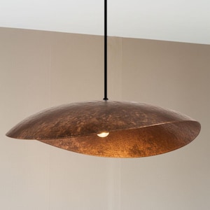 Könnte beinhalten: Eine kupferfarbene Pendelleuchte aus gehämmertem Metall. Das Licht hat eine einzigartige, geschwungene, blattartige Form mit einer einzelnen Glühbirne. Die Leuchte hängt an einem schwarzen Kabel und schafft ein modernes und künstlerisches Beleuchtungselement.