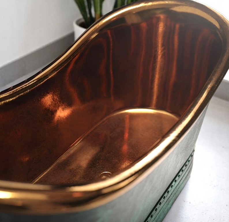 May include: A copper-colored bathtub with a gold-colored rim. The interior of the tub reflects light, creating a warm, inviting glow. The exterior has a dark green patina, adding a vintage aesthetic. The tub is a freestanding design.