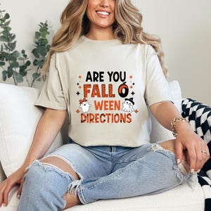 Peut inclure: T-shirt beige avec l'inscription "ARE YOU FALL O WEEN DIRECTIONS" en noir, orange et blanc. Le motif comprend une citrouille, des fantômes et des étoiles. La personne porte un jean bleu.
