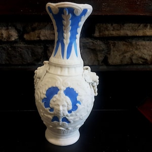 Pode incluir: Um vaso de cerâmica branco com detalhes azuis. O vaso apresenta um design em relevo detalhado com um rosto e padrões florais. A parte superior e o interior são azuis, e tem uma alça decorativa em cada lado. O vaso tem aproximadamente 20 cm de altura.