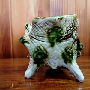 May include: A ceramic planter shaped like a tree stump, featuring a white base with green and brown accents. The planter has three legs and a textured surface, giving it a rustic appearance. The planter is on a wooden surface.
