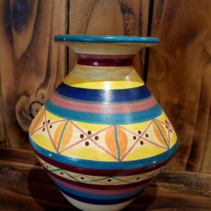 Peut inclure: Un vase en céramique peint à la main, avec une base large et un bord évasé. Le vase présente des bandes horizontales de couleurs vives, notamment jaune, bleu, rose et marron. Des motifs géométriques avec des formes de feuilles orange et des points ornent le corps du vase.