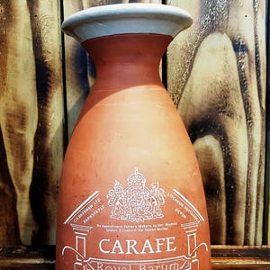 Puede incluir: Una jarra de color terracota con un borde gris claro. La jarra tiene una etiqueta con el texto "CARAFE Royal Barum Ware" y el Escudo Real. El fondo es una superficie de madera.