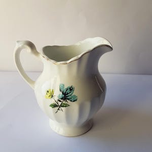 Könnte beinhalten: Weiße Keramik-Milchkanne mit geriffeltem Design und einem Blumenmuster in Blau-, Gelb- und Grüntönen.