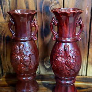Peut inclure: Une paire de vases en céramique rouge foncé assortis avec une finition brillante. Chaque vase présente deux anses latérales, un bord festonné évasé et un motif en relief sur le corps représentant une grappe de fraises avec des feuilles détaillées.