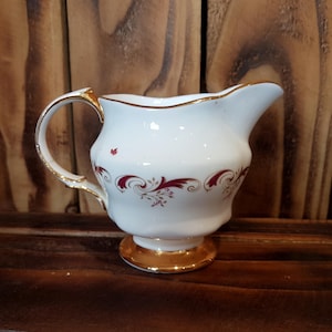 Può includere: Lattiera in ceramica bianca con manico e base dorati. La lattiera presenta un motivo floreale bordeaux e bordi dorati. La lattiera è su una superficie di legno.