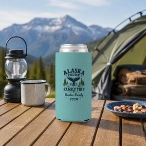 May include: A light blue foam can cooler featuring a dark blue graphic of a whale tail, pine trees, and the text "ALASKA CRUISE FAMILY TRIP Baxter Family 2026". The cylindrical sleeve is designed to hold a standard beverage can securely.