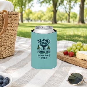 May include: A light blue foam can cooler featuring a dark blue graphic of a whale tail, pine trees, and the text "ALASKA CRUISE FAMILY TRIP", "Baxter Family", and "2026". The cylindrical sleeve is designed to insulate a standard metal beverage can.