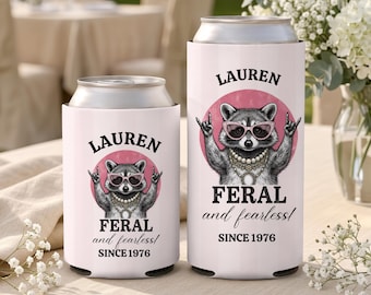 May include: Two white can coolers featuring a graphic of a raccoon wearing pink sunglasses and a pearl necklace, making a rock-and-roll hand gesture. The text reads LAUREN, FERAL and fearless!, and SINCE 1976. One cooler is short and one is tall.