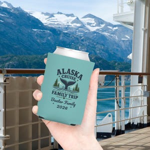 May include: A light blue foam can cooler featuring a dark blue graphic of a whale tail, pine trees, and the text ALASKA CRUISE FAMILY TRIP, Baxter Family, and 2026. The design includes small star and heart accents printed on the front surface.