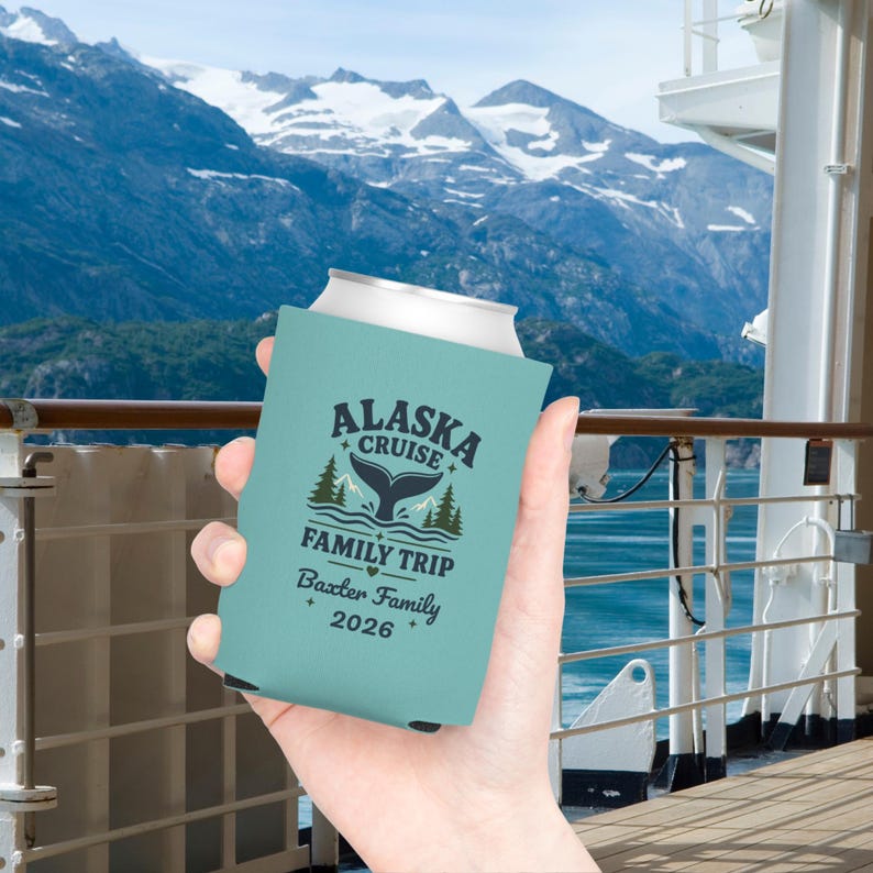 May include: A light blue foam can cooler featuring a dark blue graphic of a whale tail, pine trees, and the text ALASKA CRUISE FAMILY TRIP, Baxter Family, and 2026. The design includes small star and heart accents printed on the front surface.