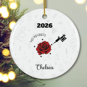 Könnte beinhalten: Ein rundes, weißes Keramikornament mit der Jahreszahl 2026 oben, dem Namen Chelsea unten und einer zentralen Grafik aus einer roten Rose, einer schwarzen Tätowiermaschine und dem Text No Regrets, umgeben von einem blassen grauen Muster.