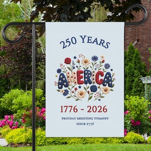 May include: A white garden flag with the text "250 YEARS AMERICA 1776-2026" in navy and red. The word "AMERICA" is decorated with floral elements in red, white, and blue. The flag is displayed on a black metal stand in a garden setting.
