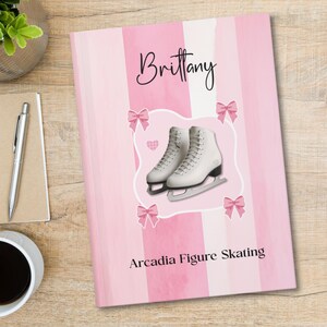 May include: A notebook with a pink and white vertical striped cover featuring the name Brittany in black script, a pair of white figure skates, four pink bows, a small pink heart, and the text Arcadia Figure Skating in black font.