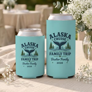 May include: Two light blue foam can coolers featuring a dark blue graphic of a whale tail, pine trees, and mountains. The text reads ALASKA CRUISE FAMILY TRIP, Baxter Family, and 2026. One cooler is short for a standard can, the other is tall.