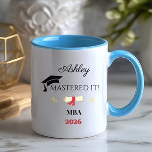 Puede incluir: Taza de cerámica blanca con interior y asa azul claro. La taza presenta el nombre "Ashley", un birrete, la frase "MASTERED IT!", un diploma y "MBA 2026" en texto rojo. Un regalo de celebración.