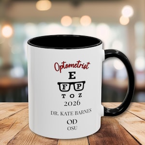 May include: White ceramic mug with a black interior and handle. The mug features the word "Optometrist" in red script, along with eye chart letters and the year 2026. The mug also includes the name "Dr. Kate Barnes, OD, OSU."