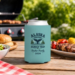 May include: A light blue foam can cooler featuring a dark blue graphic of a whale tail, pine trees, and mountains. The text reads ALASKA CRUISE FAMILY TRIP, Baxter Family, and 2026. The design includes small star accents above and below the text.