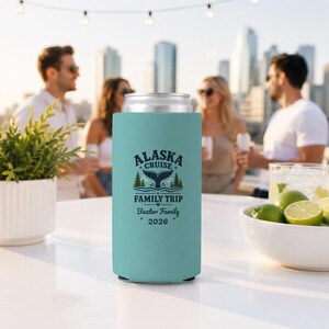 May include: A light blue foam can cooler featuring a dark blue graphic of a whale tail, pine trees, and the text ALASKA CRUISE FAMILY TRIP, Baxter Family, and 2026. The cylindrical sleeve is designed to hold a standard beverage can securely.