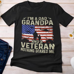 以下が含まれることがあります： アメリカの国旗、男性と子供のシルエット、そして「I'm a Dad Grandpa and a Veteran Nothing Scares Me.」というテキストが描かれた黒いTシャツです。