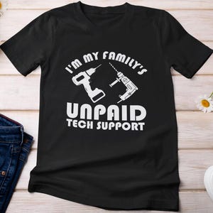 Może przedstawiać: Czarny t-shirt z białym napisem "I'M MY FAMILY'S UNPAID TECH SUPPORT" i dwiema białymi ilustracjami wiertarek. Koszulka wykonana jest z miękkiego materiału i ma krótkie rękawy.