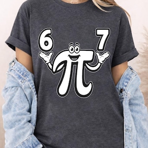 May include: Dark gray t-shirt featuring a playful design with the numbers "6" and "7" on either side of a smiling pi symbol. The pi symbol has arms and hands. The design is in white. The shirt is a casual style.