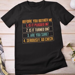 Może przedstawiać: Czarny t-shirt z napisem "BEFORE YOU BOTHER ME" i numerowaną listą kontrolną: "1. IS IT PLUGGED IN?", "2. IS IT TURNED ON?", "3. ARE YOU SURE?" i "4. SERIOUSLY, GO CHECK". Tekst jest w stylu retro.