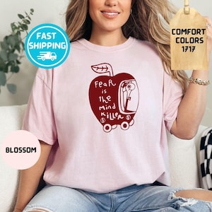 May include: A light pink t-shirt with a red apple graphic containing the text "Fear is the mind killer." The shirt also has a "Fast Shipping" badge and a "Blossom" color tag. The Comfort Colors 1717 tag is also visible.