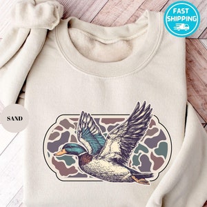 Peut inclure: Un sweat-shirt beige avec un imprim&eacute; graphique d'un canard volant au-dessus d'un motif camouflage. Le texte "SAND" est imprim&eacute; sur un cercle blanc.