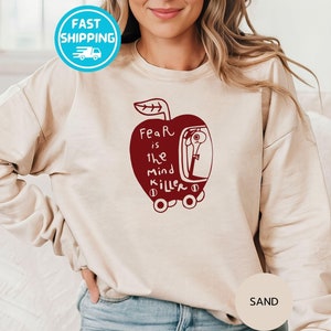 May include: A beige sweatshirt with a red apple graphic. The apple has a face and the text "Fear is the mind killer" inside. The text "SAND" is on the bottom right corner of the image.
