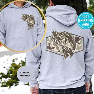 Op de afbeelding: Atletische heather grijze hoodie met een voorzak en een capuchon. De hoodie heeft een visafbeelding op de voor- en achterkant. De achterste afbeelding is een vis in een bruine en groene camouflage zeshoek.