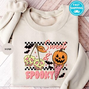 Op de afbeelding: Zandkleurige sweatshirt met een Halloween-thema. Het ontwerp toont een pompoen-ijshoorntje, schedelkersen en vleermuizen, met de woorden "Sweet & Spooky" in roze. Een scrunchie is zichtbaar op de linkerschouder.