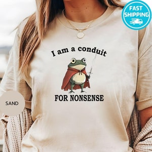 May include: A beige t-shirt with the text "I am a conduit for nonsense" and a cartoon frog wearing a red cape and holding a sword.