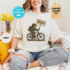 May include: Ivory-colored t-shirt featuring a whimsical illustration of a frog riding a bicycle and holding a sign that reads "RESIST." The frog wears a hat and backpack. The shirt has a Comfort Colors 1717 tag.