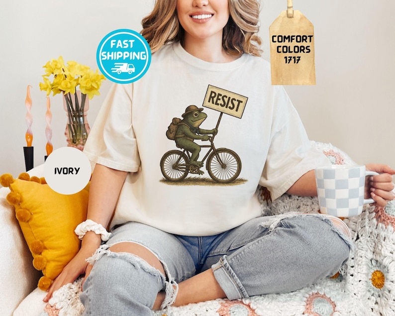 May include: Ivory-colored t-shirt featuring a whimsical illustration of a frog riding a bicycle and holding a sign that reads "RESIST." The frog wears a hat and backpack. The shirt has a Comfort Colors 1717 tag.