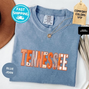 May include: A blue jean Comfort Colors t-shirt with the word "TENNESSEE" embroidered in orange and patterned fabric. The shirt has a round neckline and short sleeves. A gold necklace and a tag that says "COMFORT COLORS 1717" are also visible.