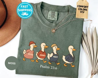 May include: A moss green t-shirt featuring a graphic of four ducks walking in a line. The ducks wear clothing with the text Shirley, Goodness, and Mercy printed on their shirts. Below the ducks, the text Psalm 23:6 is printed in white.