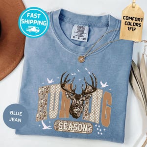 May include: Pale blue t-shirt featuring a deer graphic and the words "Hunting Season". The design uses brown and tan tones. The shirt has a Comfort Colors tag and a Fast Shipping sticker.