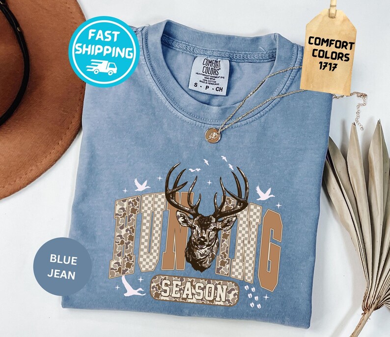 May include: Pale blue t-shirt featuring a deer graphic and the words "Hunting Season". The design uses brown and tan tones. The shirt has a Comfort Colors tag and a Fast Shipping sticker.