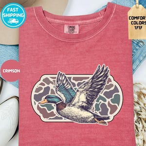 May include: Crimson-colored t-shirt featuring a detailed illustration of a duck in flight, set against a camouflage-style background. The shirt has a crew neck and short sleeves. The tag reads "Comfort Colors 1717".